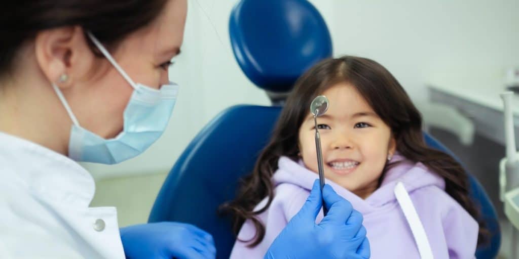 Dentist wearing mask showing dental mirror to young patient.